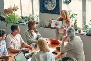 Femme présentant un graphique coloré à un groupe assis autour d'une table dans une salle lumineuse.