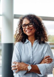 Femme souriante en chemise rayée, lunettes et montre, bras croisés dans un espace lumineux.