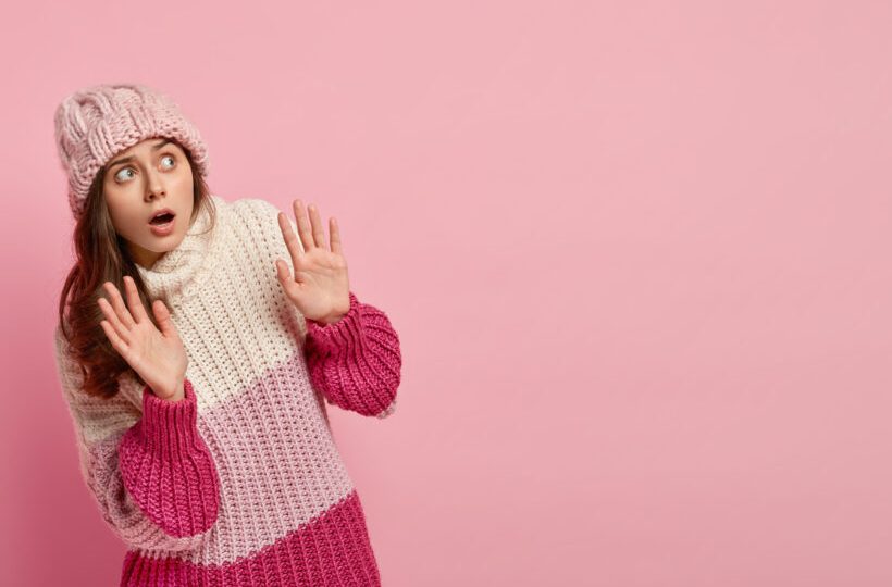 Shot of scared frightened young woman keeps hands in protective gesture, looks upwards, screams from fear, dressed in warm winter jumper and knitted headgear, isolated over pink studio wall.